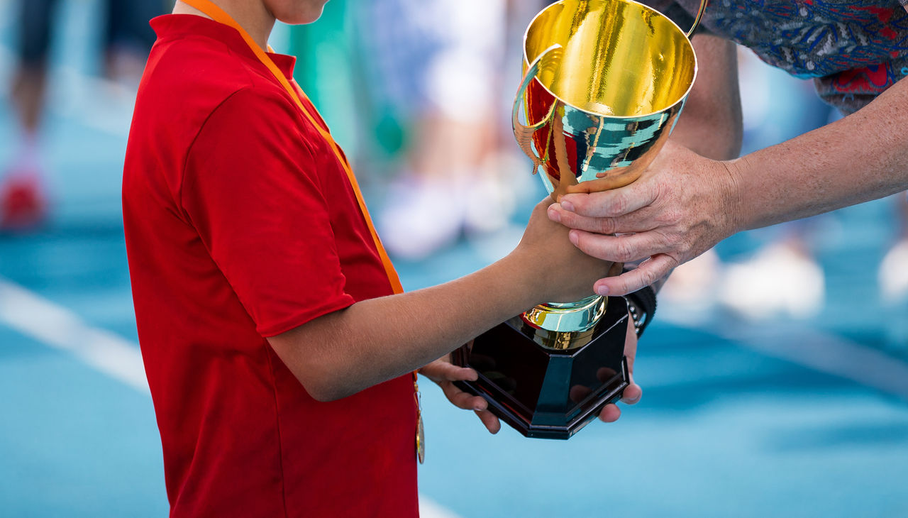 Child in a sportswear receiving a golden cup. Young athlete winning the sports school competition. Boy with golden medal getting an award for the best player of the tournament