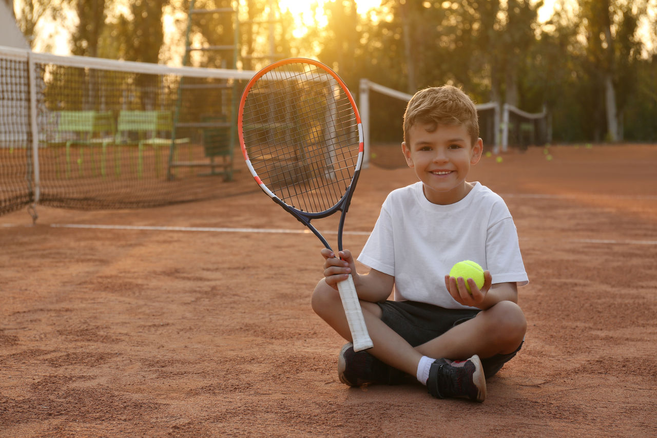 Cute little boy with tennis racket and ball on court outdoors