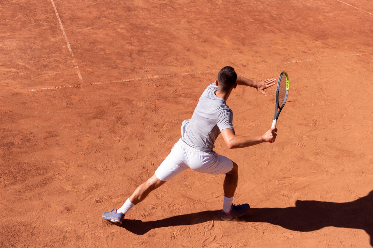 Professional tennis player performs forehand hit on clay tennis court. Young male athlete with tennis racket in action. Junior tennis sport. Back view, shadow, copy space