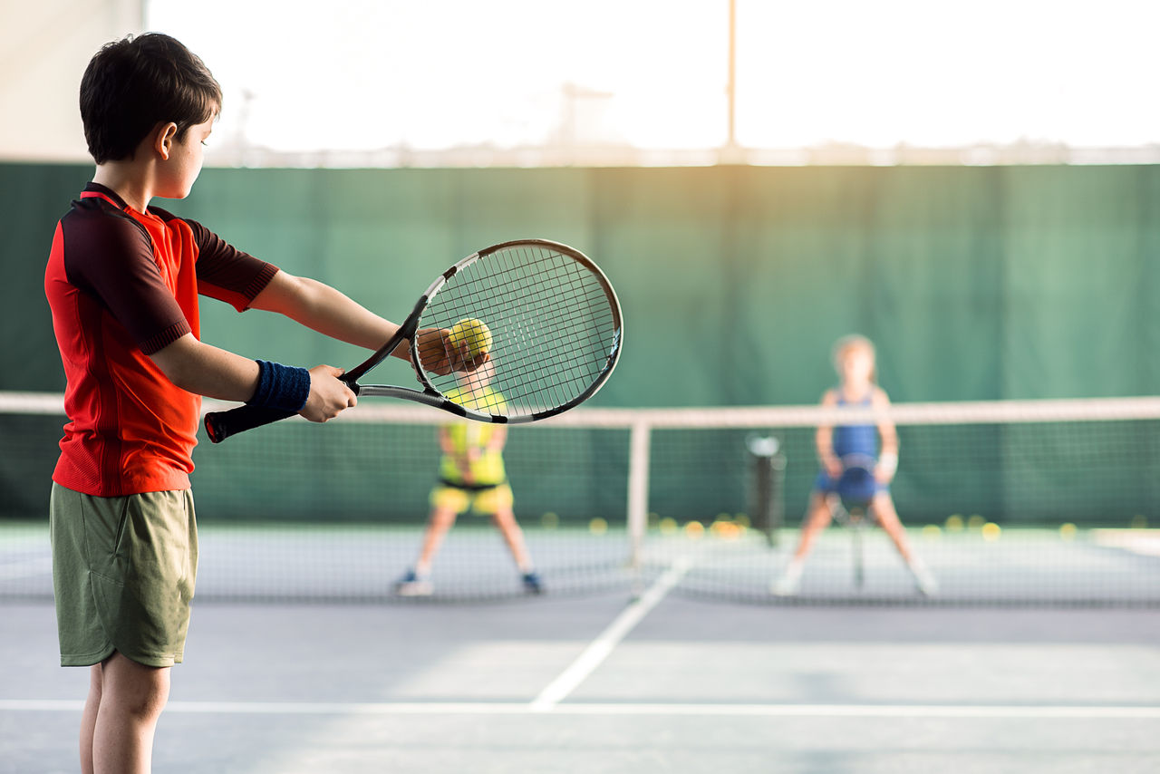 Concentrated boy is pitching tennis ball to children while raising racket. Copy space in right side