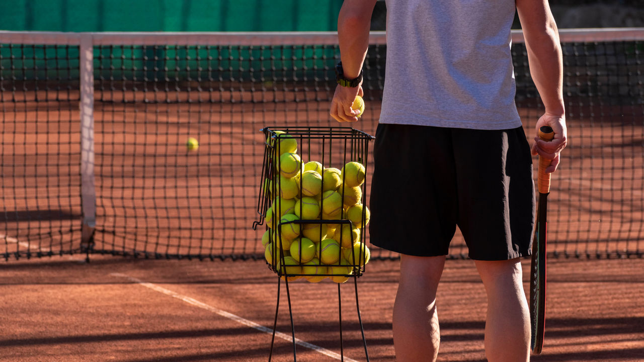 Tennis coach conducts training on red clay court, basket with tennis balls near him. Blurred background. Sports activity and leisure concept. Lifestyle