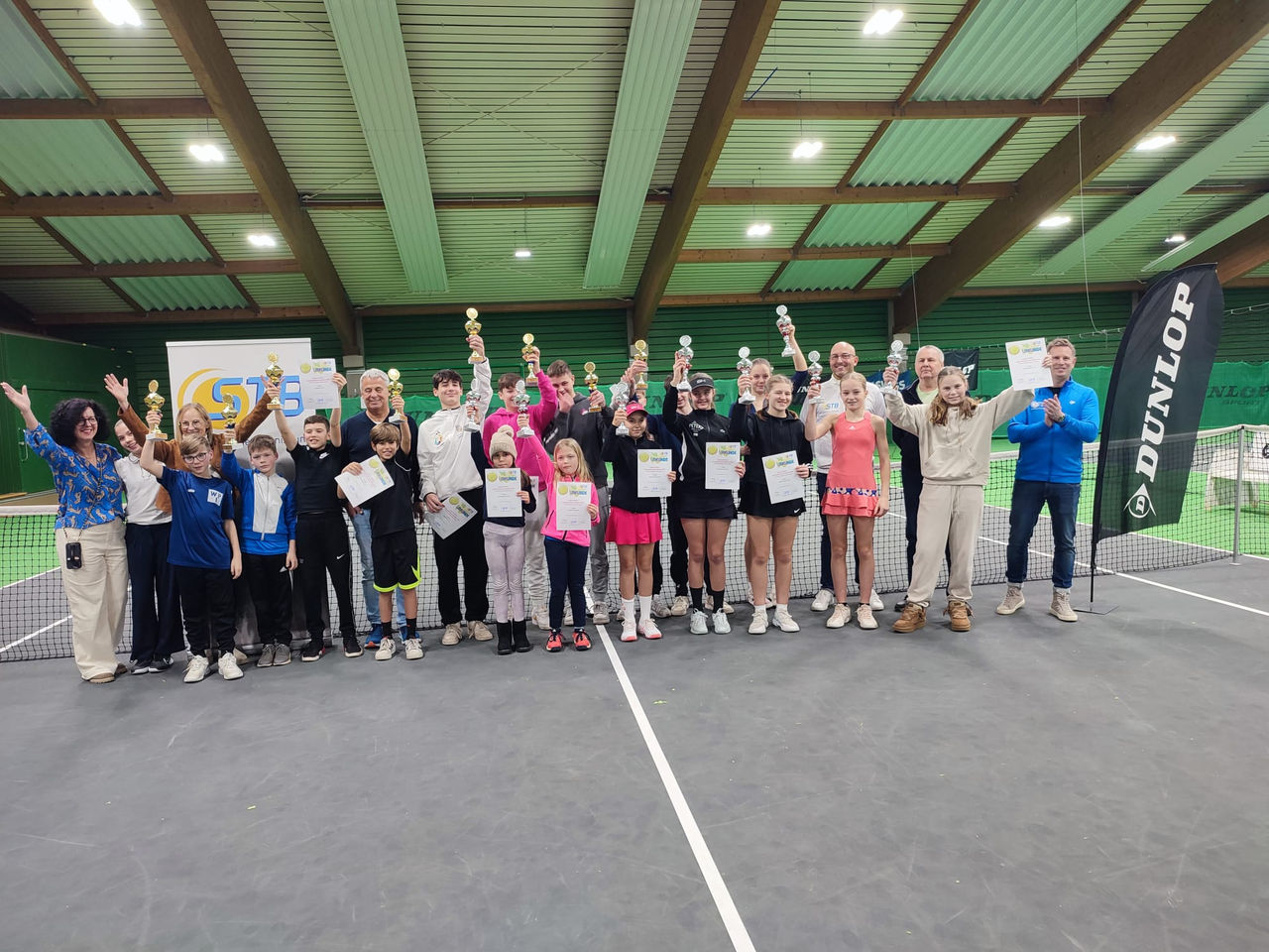 Alle Sieger:innen des STB-Kaders jubelnd vor dem Netz auf dem Platz in der Halle mit Pokalen und Urkunden