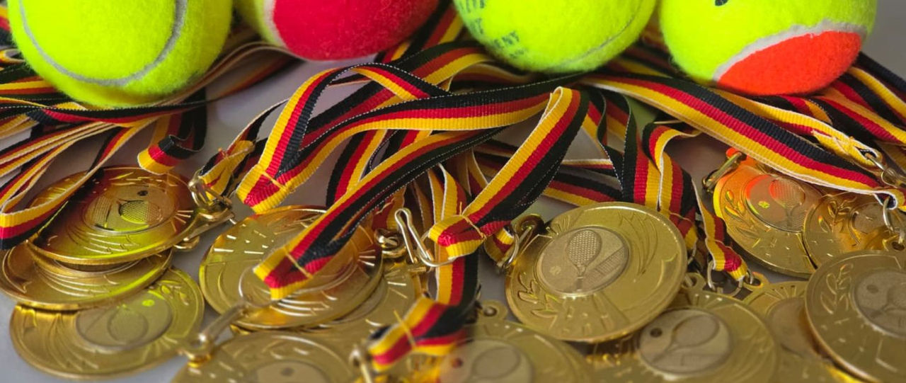 Goldene Medaillen mit einem Tennisschläger drauf und  verschiedene Sorten Tennisbälle liegend auf dem Tisch