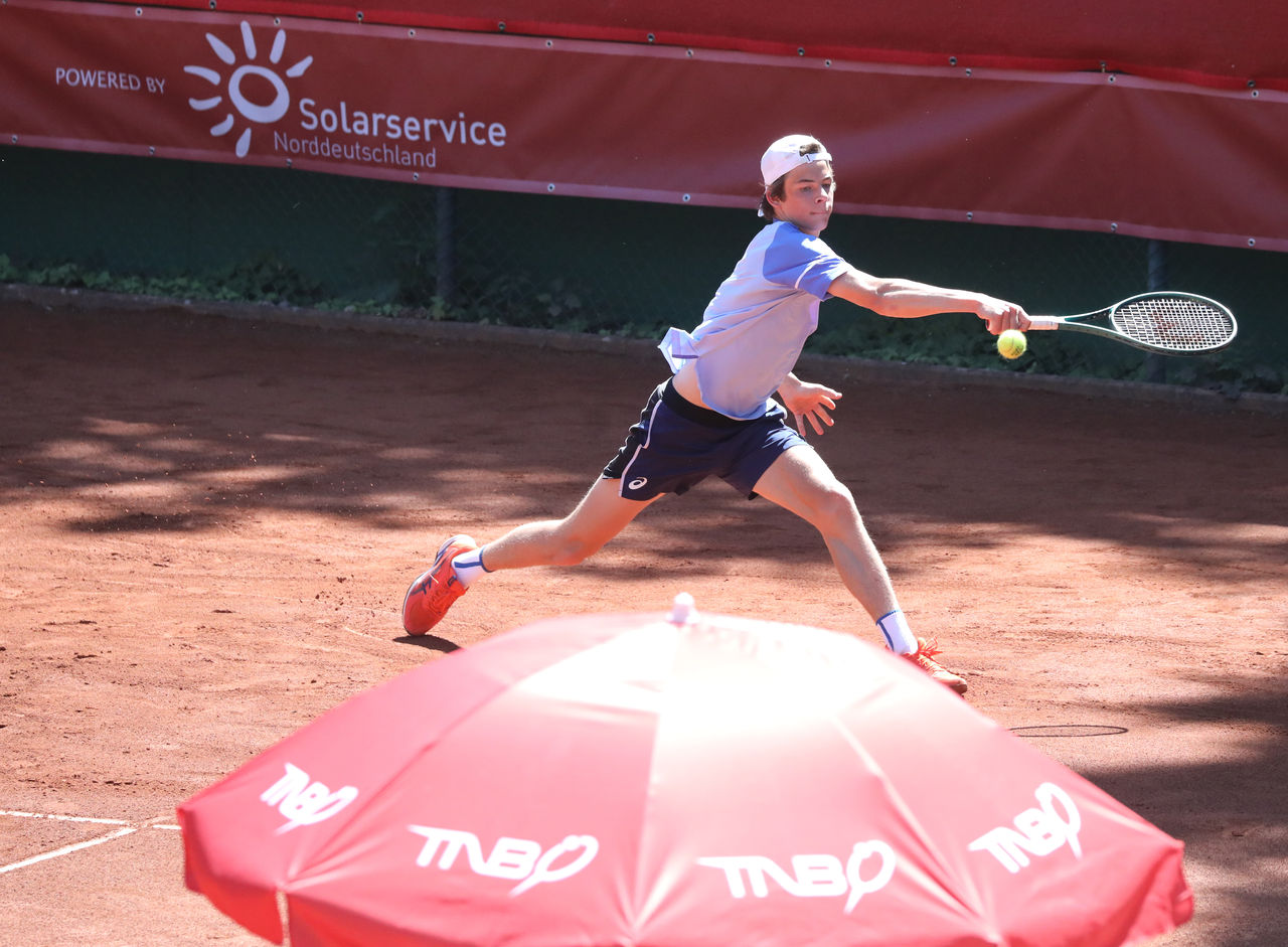  Ein Tennisspieler auf einem Sandplatz schlägt einen Ball. Im Vordergrund ein roter Sonnenschirm mit TNB-Logo, im Hintergrund ein Werbebanner von „Solarservice Norddeutschland“.