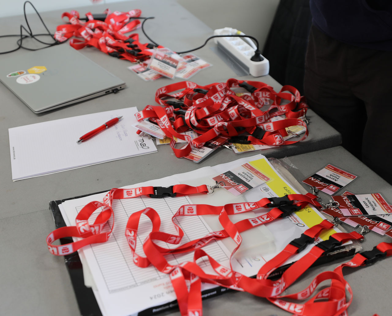 Ein Tisch mit roten Lanyards, Namensschildern, Notizblöcken, einem Laptop und einer Mehrfachsteckdose, vorbereitet für eine Veranstaltung.