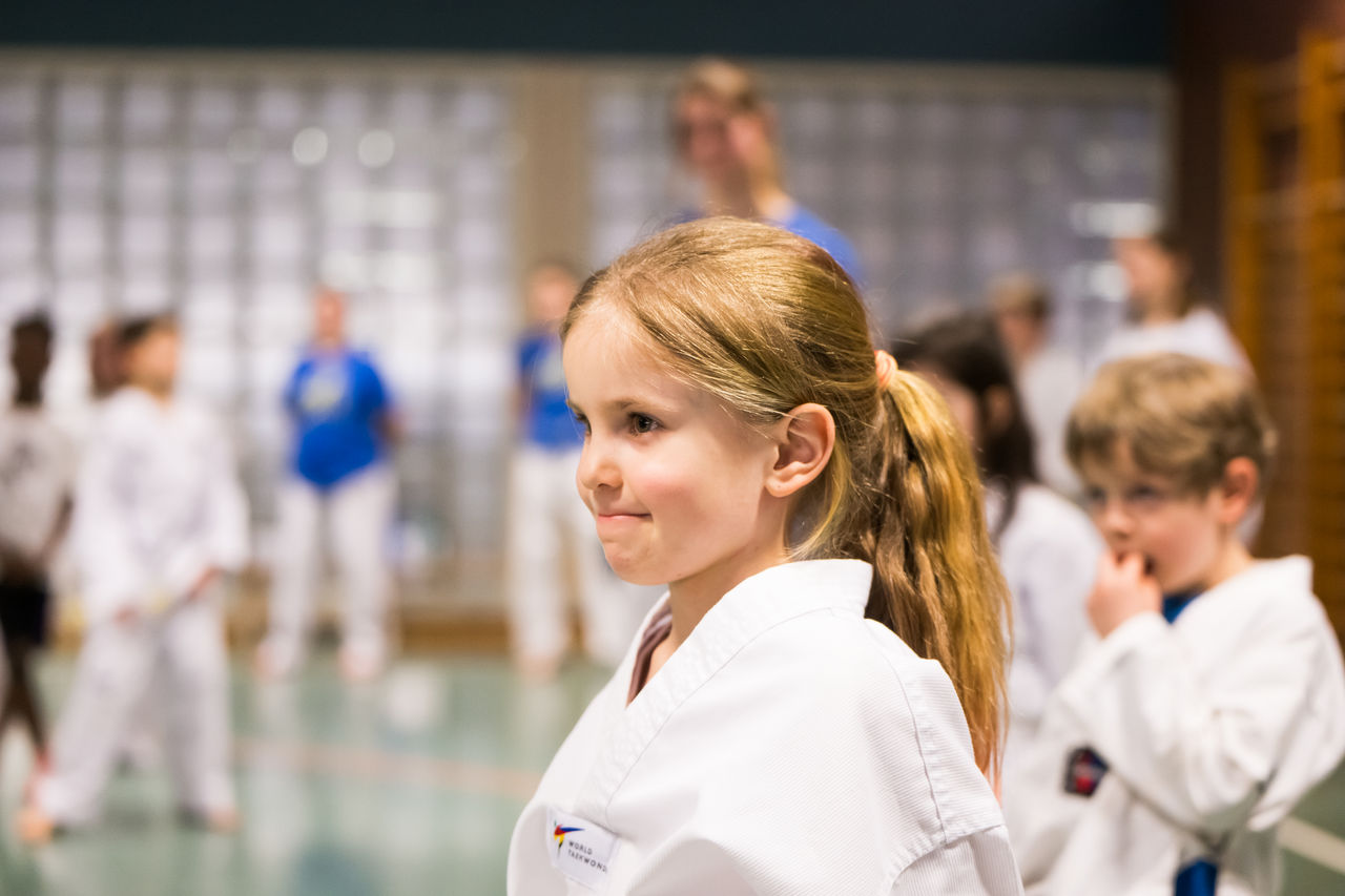 Mädchen in weißem Karateanzug steht in Sporthalle, weitere Kinder in Kampfsportkleidung im Hintergrund.